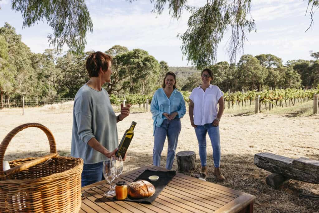 Three women enjoying a relaxed wine tasting at a Margaret River vineyard, chatting under shady trees beside rows of vines.
