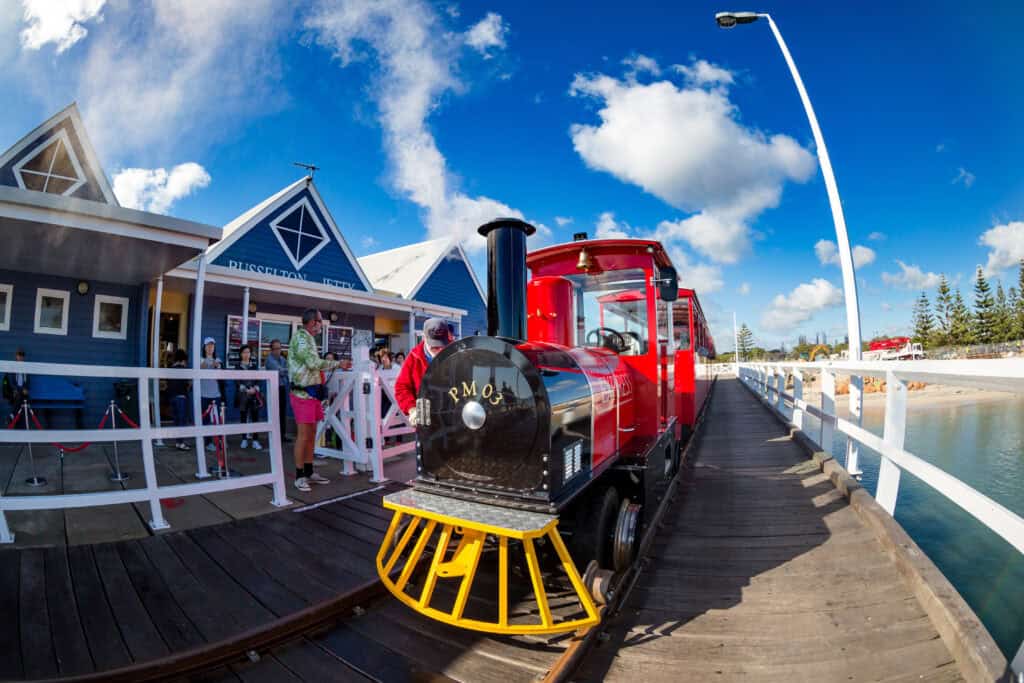 Wide-angle photograph of the historic red steam engine on the Busselton Jetty, positioned on the timber boardwalk next to the passenger boarding area and blue buildings.